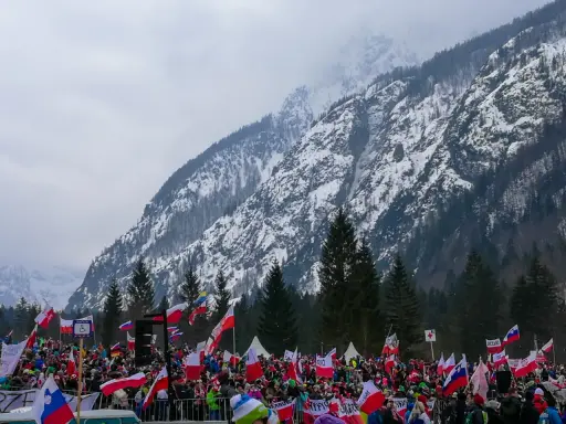 Tłum kibiców z flagami na tle ośnieżonej góry, atmosfera na skoczni w Planicy.