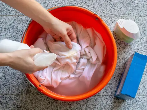 Ręce rozpylają płyn na poplamioną tkaninę w pomarańczowym wiadrze z wodą.