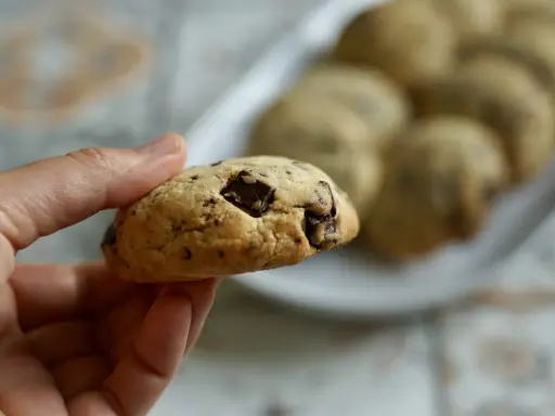 Une main tient un cookie épais et moelleux aux pépites de chocolat. D'autres cookies sont disposés sur une assiette en arrière-plan.
