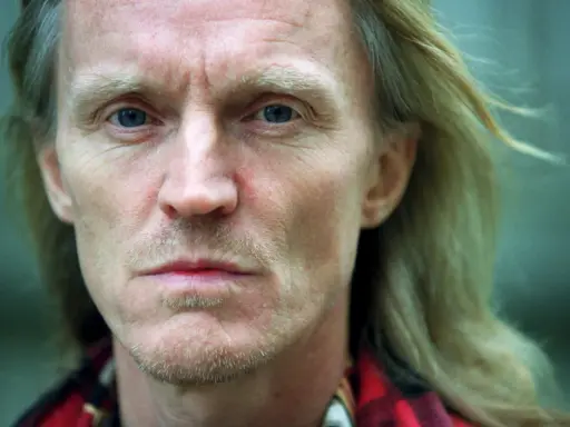 Närbild på John Holm med blå ögon och blont hår. Hans ansikte bär spår av livserfarenhet, en sordinerad blick.