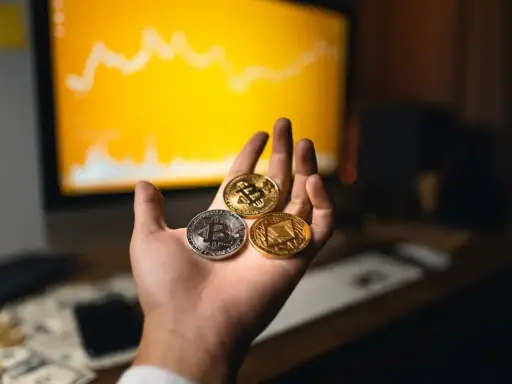 Dłoń trzyma trzy monety kryptowalut: Bitcoin, Ethereum i inną. W tle wykres na monitorze, sugerujący handel na kantory kryptowalut.