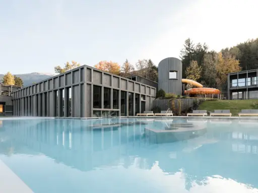 Piscina esterna con acqua fumante e scivoli acquatici arancioni al Aki Family Resort Plose. Architettura moderna in legno e montagne sullo sfondo.