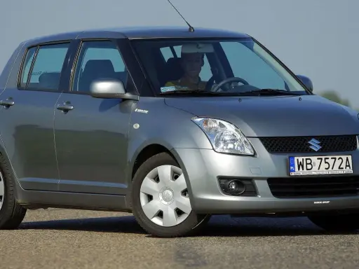 Szary Suzuki Swift 2007 z kierowcą za kierownicą, zaparkowany na drodze.