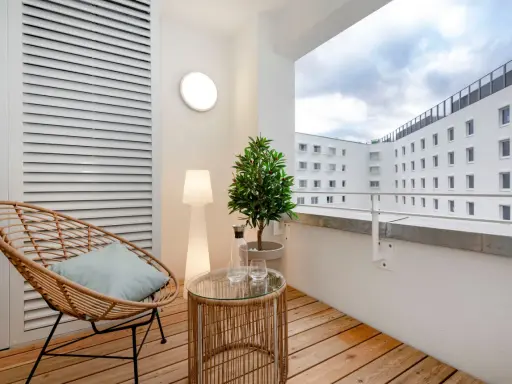 Przytulny balkon z drewna, z wiklinowym fotelem, stolikiem i rośliną. W tle białe budynki.