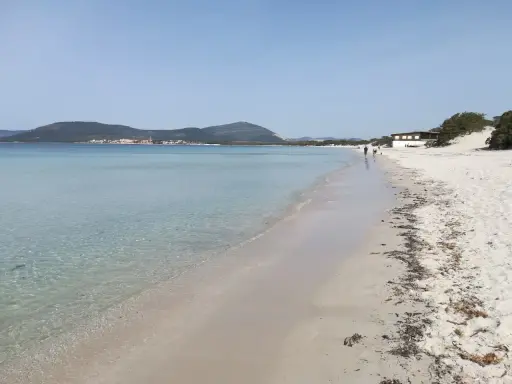 Spiaggia della Fertilia con acque cristalline, sabbia bianca e un edificio in lontananza. Due persone passeggiano lungo la riva.