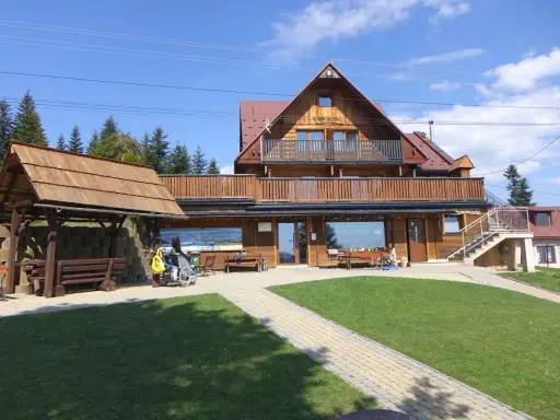 Drewniana bacówka na obidzy z balkonem i tarasem, otoczona zielenią i ścieżkami. Obok wózek inwalidzki z żółtą torbą.