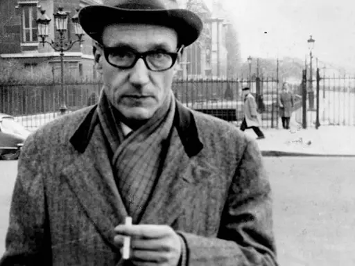 William Burroughs, con cappello e occhiali, tiene una sigaretta in mano, passeggiando in una strada urbana con persone sullo sfondo.