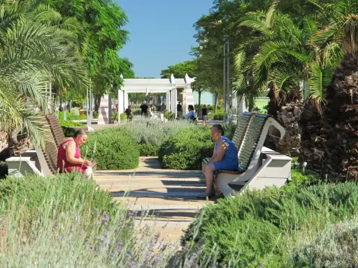Due donne sedute su panchine in un parco curato, con palme e vegetazione rigogliosa, in una giornata di sole a Riccione centro.