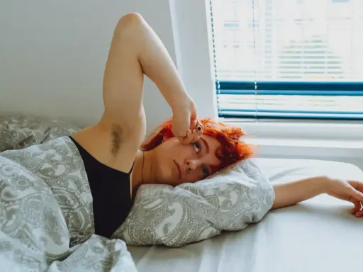 Jeune femme aux cheveux roux allongée dans son lit, le bras levé, le regard perdu. Elle semble fatiguée, peut-être un des symptômes manque magnésium.