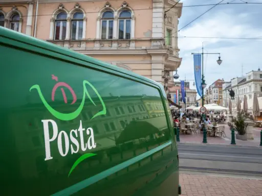 Zielona furgonetka pocztowa z logo "Posta" stoi na ulicy w węgierskim mieście. W tle widać kawiarnie i zabytkowe budynki.