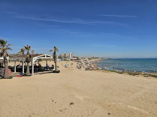 Sonniger Tag am Playa de la Mata. Menschen entspannen sich am Strand, einige im Wasser, andere unter Sonnenschirmen. Palmen säumen den Sand.
