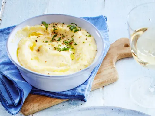 Cremige Polenta, verfeinert mit Butter und Kräutern, serviert in einer Schüssel. Ein Glas Weißwein und ein Holzbrett vervollständigen dieses Polenta Rezept.