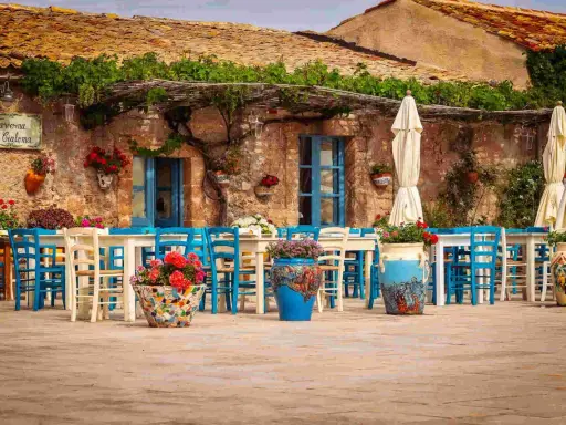 Taverna La Cialoma, un angolo incantevole di Siracusa e dintorni, con tavoli apparecchiati e sedie blu sotto un pergolato fiorito.