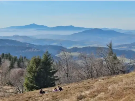 Widok na Beskid Wyspowy z kilkoma osobami odpoczywającymi na zboczu. W oddali widać kolejne pasma górskie w lekkiej mgle.