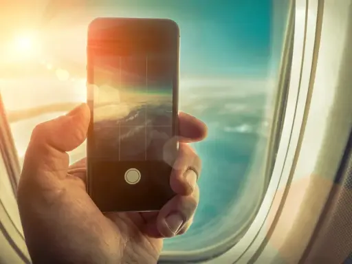 Dłoń trzyma smartfon, fotografując chmury z okna samolotu. Czy w samolocie można używać telefonu? Tak, do robienia zdjęć!
