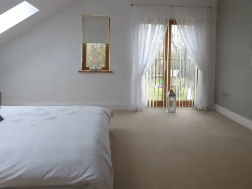 Dormitorio con cama blanca, ventana abuhardillada y puerta acristalada con cortinas. La orientación oeste de los pisos permite una luz suave.