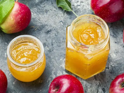 Zwei Gläser mit leuchtend gelbem Apfelgelee, umgeben von frischen roten Äpfeln. Ein köstliches Apfelgelee Rezept zum Nachmachen.