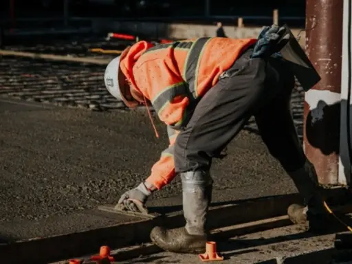 Obrero con casco y chaleco naranja trabajando con hormigón autocompactante, alisando la superficie con una llana.
