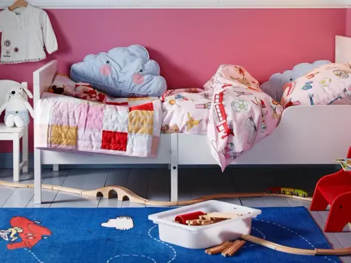 Habitación infantil con dos camas blancas, cojines de nube y mantas coloridas. Alfombra azul con tren de juguete y vías.