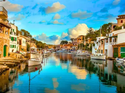 Un pintoresco pueblo pesquero con casas blancas y verdes, barcos amarrados en un canal de aguas cristalinas y un cielo azul con nubes esponjosas.