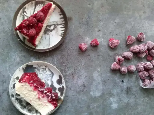 Dwa kawałki sernika z malinami na talerzach, obok garść mrożonych malin. Idealny deser na letnie popołudnie.