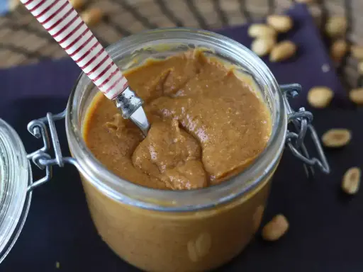 Un pot de praliné cacahuète onctueux, prêt à être dégusté. Quelques cacahuètes parsèment le plan de travail.