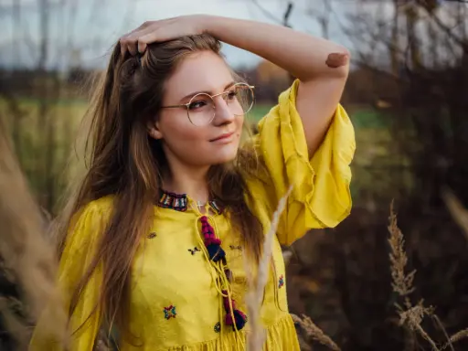 Portret dziewczyny w okularach i żółtej sukience, wśród traw. Różne rodzaje portretów w fotografii mogą uchwycić taką naturalną, swobodną chwilę.