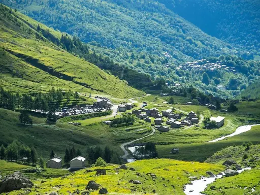 Un villaggio alpino nel **Pian della Regina**, con case in pietra e un fiume che scorre tra prati verdi e boschi lussureggianti.