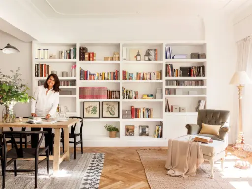 Una diseñadora casa sonríe junto a una mesa de comedor de madera, rodeada de estanterías llenas de libros y objetos decorativos.