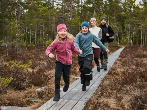 Familj njuter av aktiviteter i skogen med barn på en spång. Två barn springer glatt framför sina föräldrar i en vacker skogsmiljö.