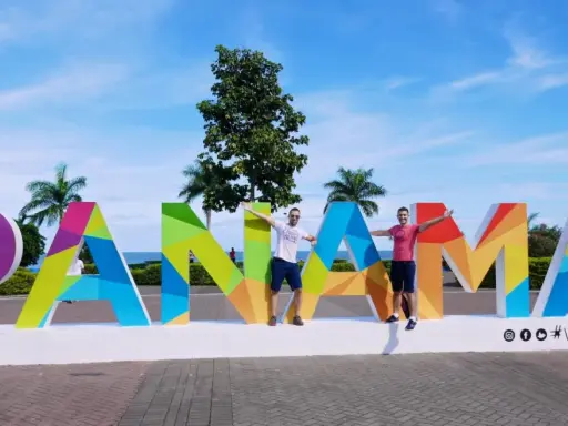 Two men pose with arms outstretched in front of a colorful "PANAMÁ" sign, suggesting Panama is gay friendly and a welcoming destination.