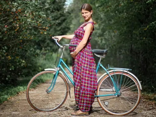 Kobieta w ciąży w kratowanej sukience opiera się o turkusowy rower na leśnej ścieżce.