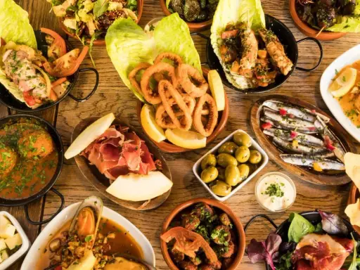 Vielfältige Tapas ideen: Tintenfischringe, Sardinen, Garnelen, Fleischbällchen, Schinken mit Melone, Oliven und mehr auf Holztisch.