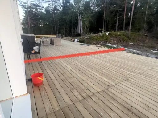 Nybyggd altan med en röd markering som visar var ett vindskydd ska byggas. Trädgårdsmöbler och en parasoll finns på plats.