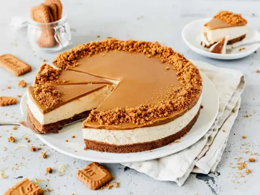 Ein köstlicher Lotus Cheesecake mit Karamellglasur und Keksbröseln. Ein Stück wurde bereits entnommen, daneben liegen weitere Kekse.