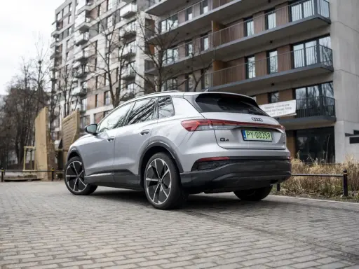 Srebrny Audi Q4 e-tron zaparkowany na brukowanym podjeździe przed nowoczesnym budynkiem mieszkalnym.