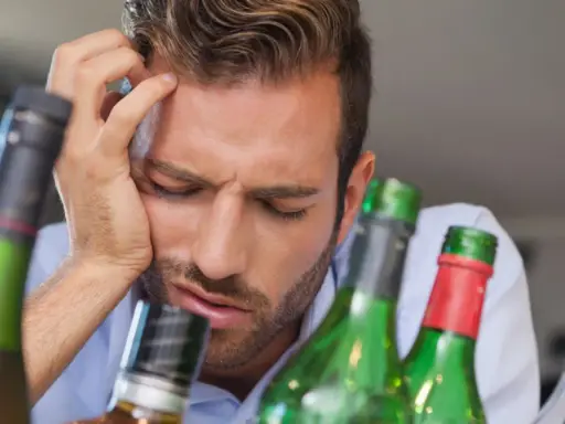 Mężczyzna z ręką na czole, otoczony butelkami, wyraźnie cierpi z powodu objawów przepicia alkoholowego.