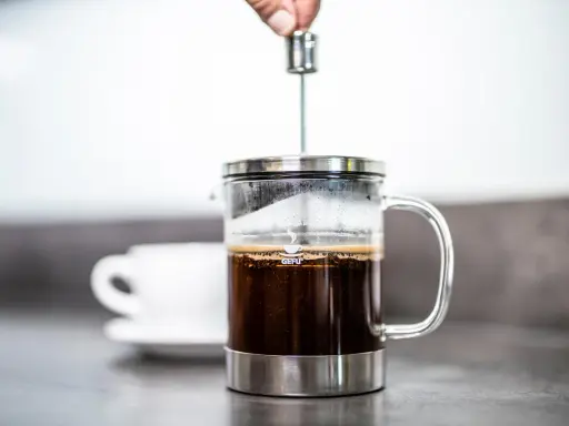 Eine Hand drückt den Stempel in eine French Press, um den perfekten Kaffee-Brew zuzubereiten. Eine Tasse wartet im Hintergrund.