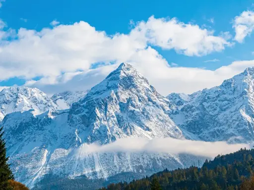 Majestatyczne, ośnieżone austria góry pod błękitnym niebem z chmurami. W dole las iglasty.