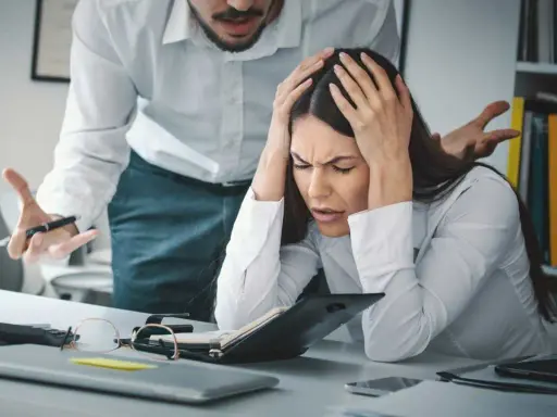 Kobieta z rękami na głowie, przytłoczona stresem w pracy. Jej szef gestykuluje, co potęguje jej cierpienie. To moment, gdy rezygnacja z pracy z powodu stresu wydaje się jedynym wyjściem.