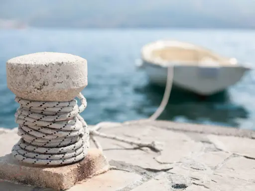 Gros plan sur un bollard en pierre avec une corde enroulée, prêt pour l'attache bateau pour quai. Un petit bateau est amarré en arrière-plan.