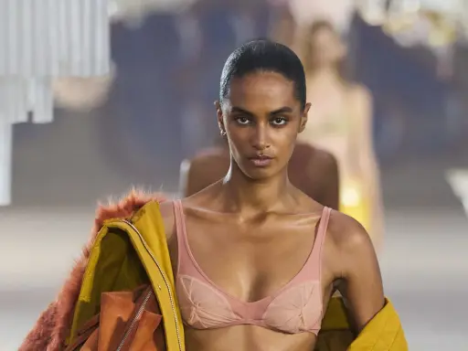 Modella in passerella con un audace reggiseno a vista color pesca, abbinato a una giacca gialla con pelliccia arancione.