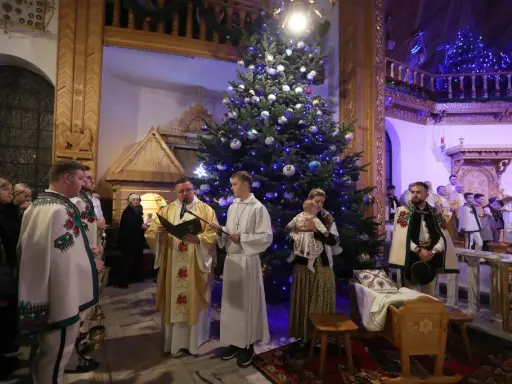 Pasterka w kościele w Zakopanem. Ksiądz czyta z księgi przy choince, obok ministranci i wierni w tradycyjnych strojach.