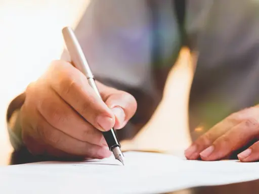 Dłoń pisze piórem po papierze, przygotowując dokument. Dowiedz się, jak napisać testament.