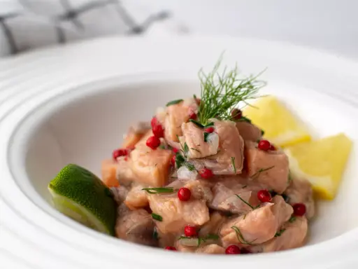Un classique tartare de saumon, frais et coloré, garni d'aneth, de baies roses et servi avec des quartiers de citron vert et de citron jaune.