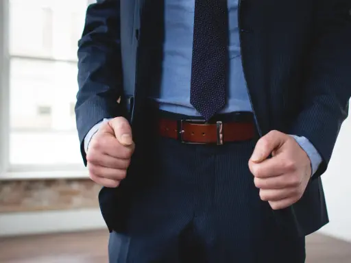 A man in a navy pinstripe suit and blue shirt wears a brown belt with black pants.