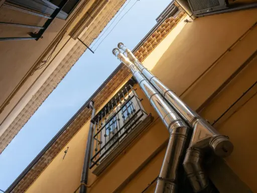 Dos tubos de salida humos estufa pellets brillan bajo el sol, ascendiendo por una pared amarilla junto a una ventana enrejada.