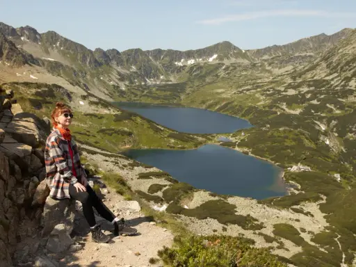 Kobieta odpoczywa na szlaku w Dolinie Pięciu Stawów. W tle rozciągają się góry i jeziora.