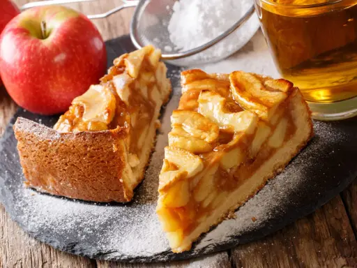 Deux parts de tarte aux pommes sur une ardoise, saupoudrées de sucre glace, avec des pommes et un verre de cidre.