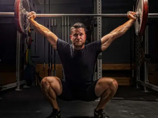 Ein Mann im tiefen Squat mit einer Langhantel über dem Kopf. Seine Muskeln sind angespannt, bereit für den nächsten Schritt im **power lifting**.